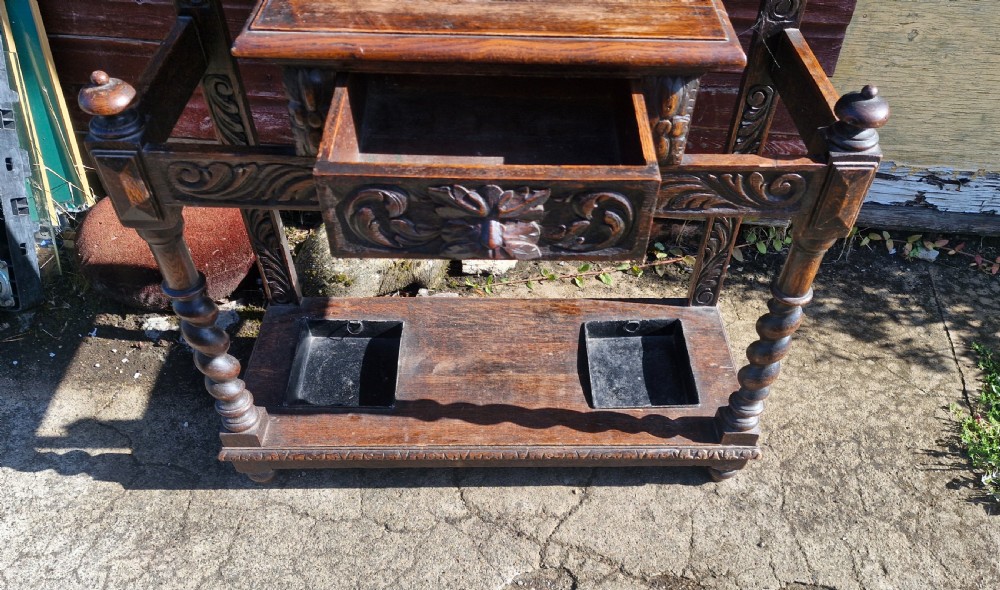A tall Victorian carved dark oak hall stand featuring Green Man mask carvings, turned pegs, a small central shelf, and lower umbrella racks with metal drip trays. View II