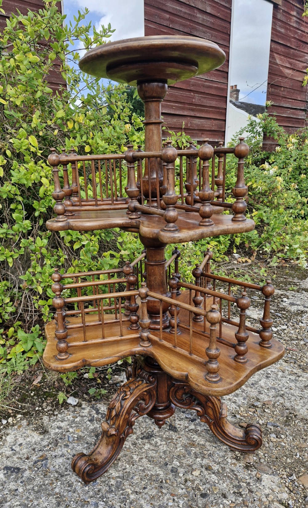 A nineteenth century revolving wooden booktable featuring tiered shelves with spindle galleries, a central turned column, and an ornate carved base.
