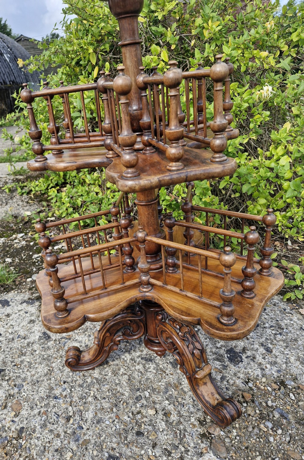 A nineteenth century revolving wooden booktable featuring tiered shelves with spindle galleries, a central turned column, and an ornate carved base. View II