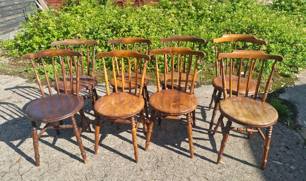 A set of eight matched antique wooden kitchen chairs featuring circular seats, spindle backs, curved crest rails, and turned legs.