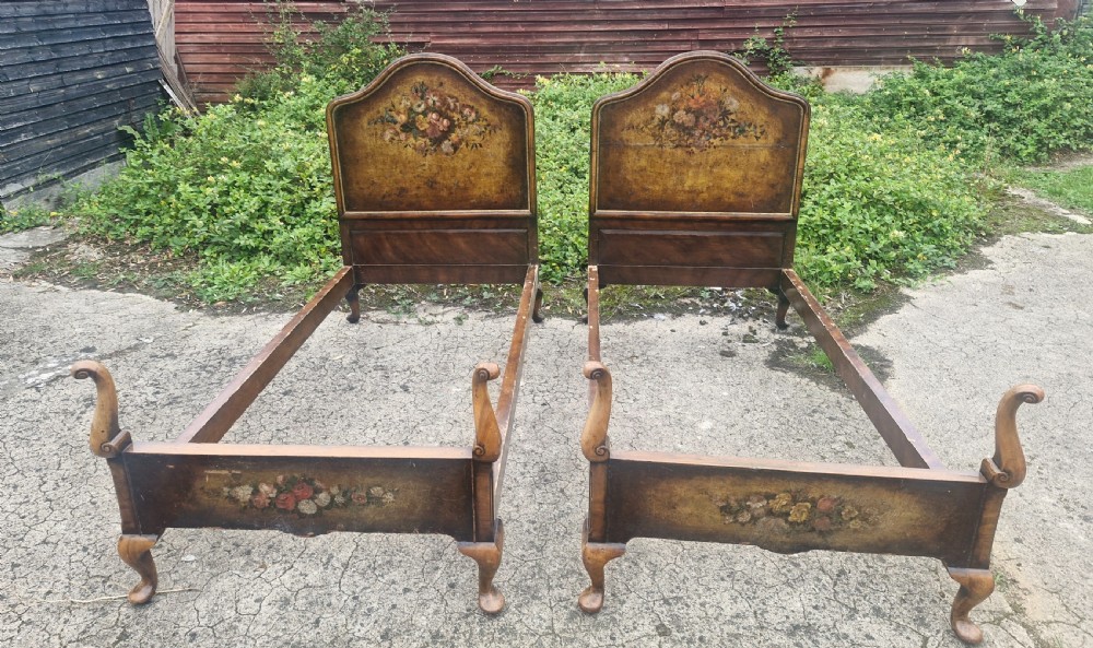 A pair of antique walnut single bed frames featuring arched headboards with floral painted leather panels and scrolled footboard posts.