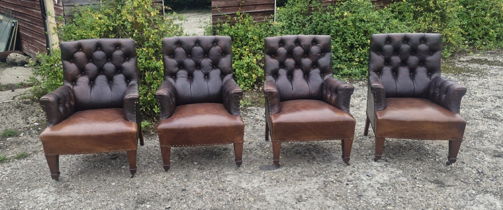 Set of four dark brown leather armchairs featuring deep button tufting, brass studding, and square tapered legs with castors.