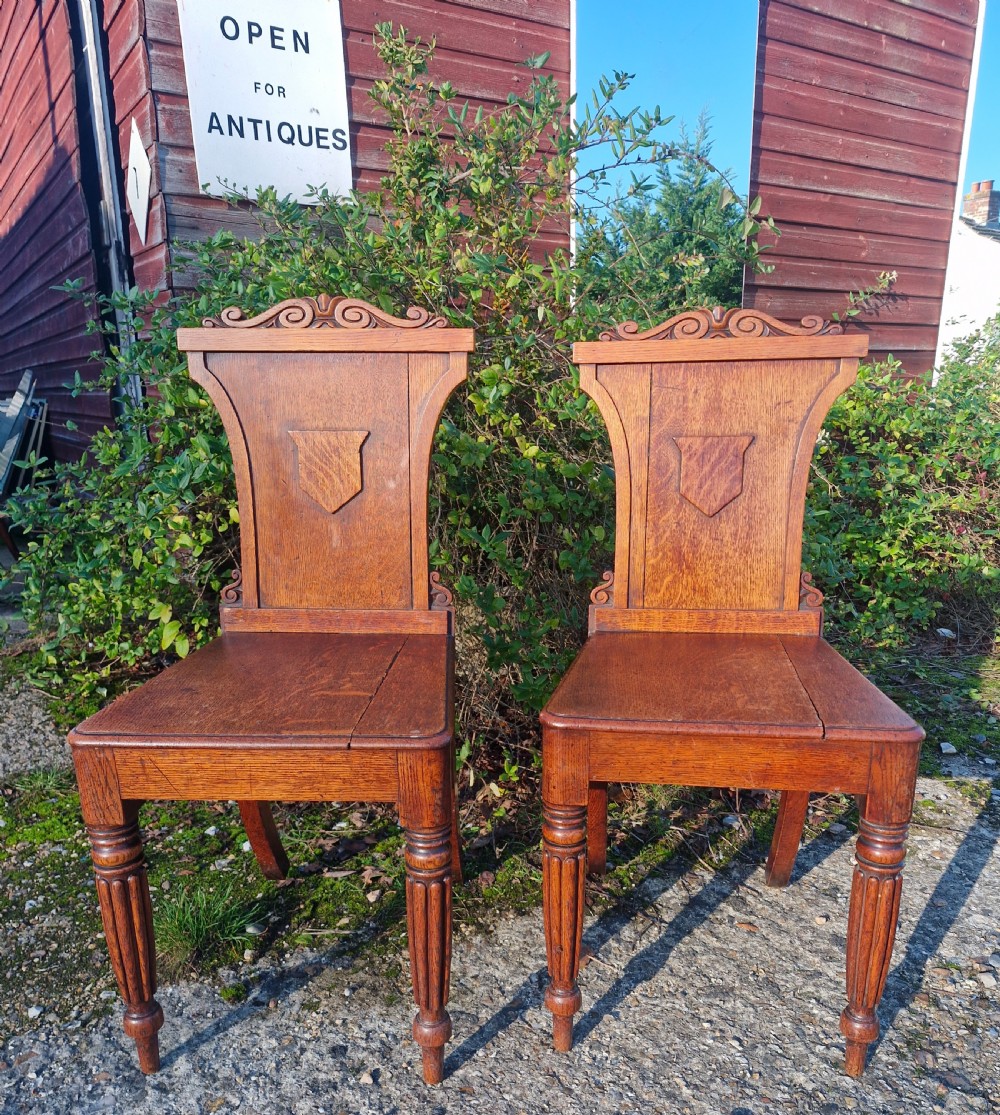 A pair of Regency oak hall chairs featuring carved scroll cresting, central shield back panels, flat wooden seats, and turned reeded front legs.