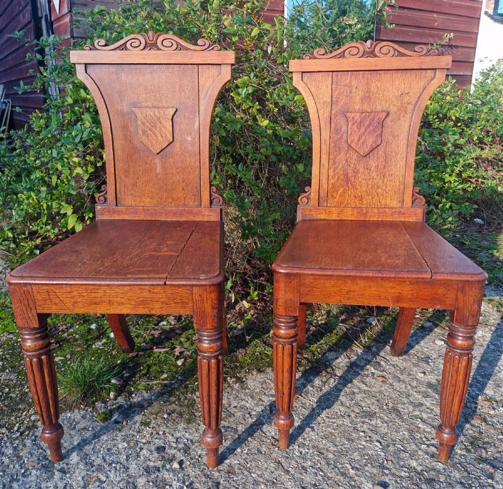 A pair of Regency oak hall chairs featuring carved scroll cresting, central shield back panels, flat wooden seats, and turned reeded front legs. View VIII