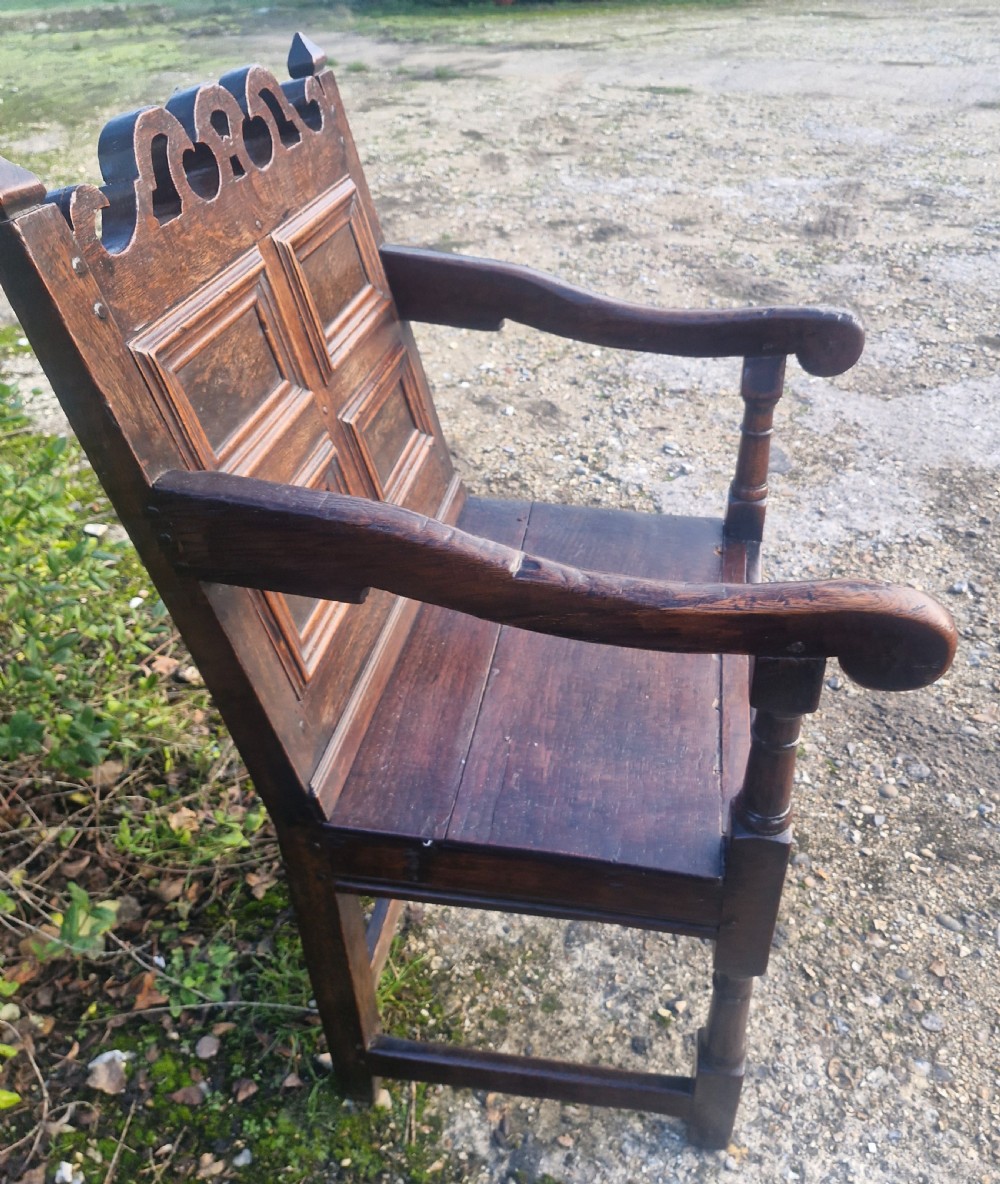 An early 18th-century dark oak wainscot armchair featuring a pierced carved cresting rail, four recessed geometric back panels, curved armrests, and turned front legs. View III
