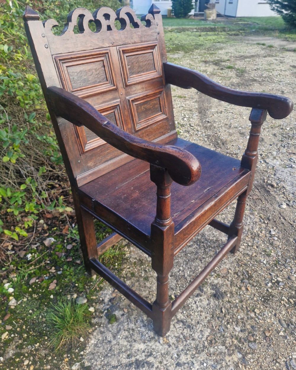 An early 18th-century dark oak wainscot armchair featuring a pierced carved cresting rail, four recessed geometric back panels, curved armrests, and turned front legs. View V