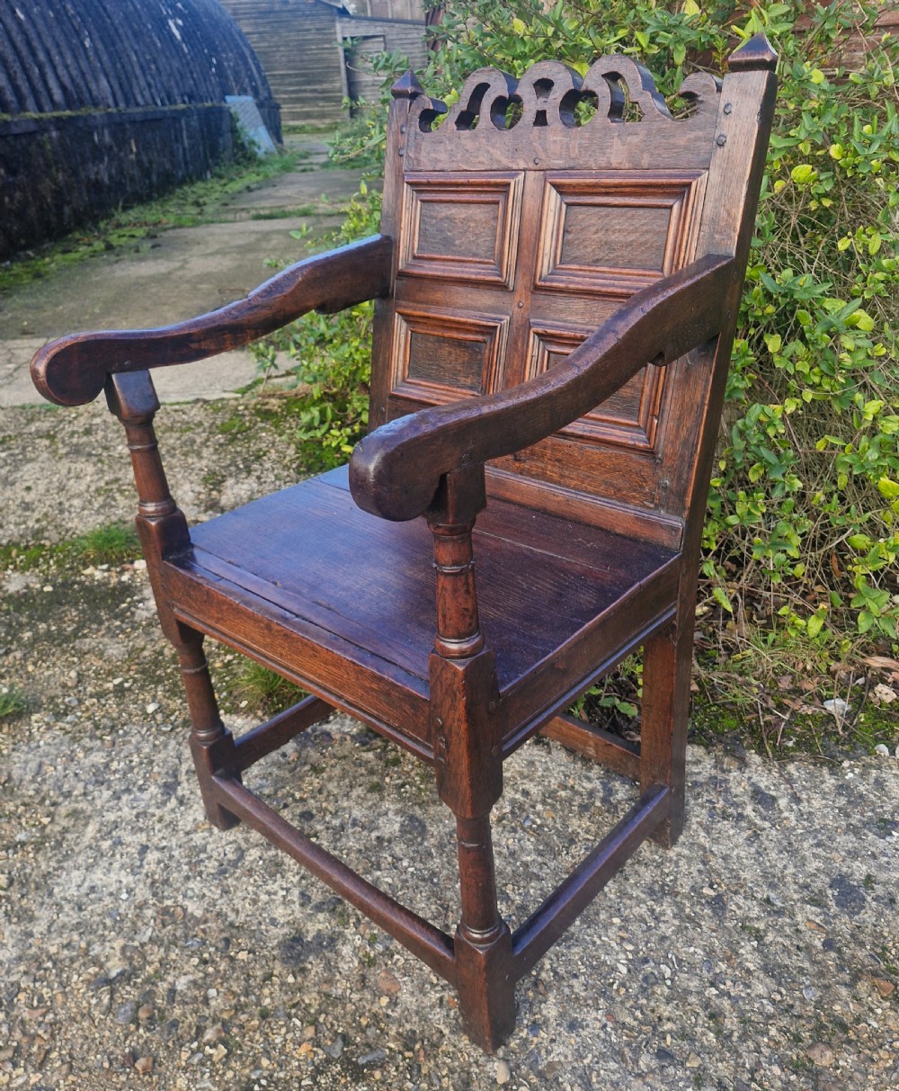 An early 18th-century dark oak wainscot armchair featuring a pierced carved cresting rail, four recessed geometric back panels, curved armrests, and turned front legs. View VI