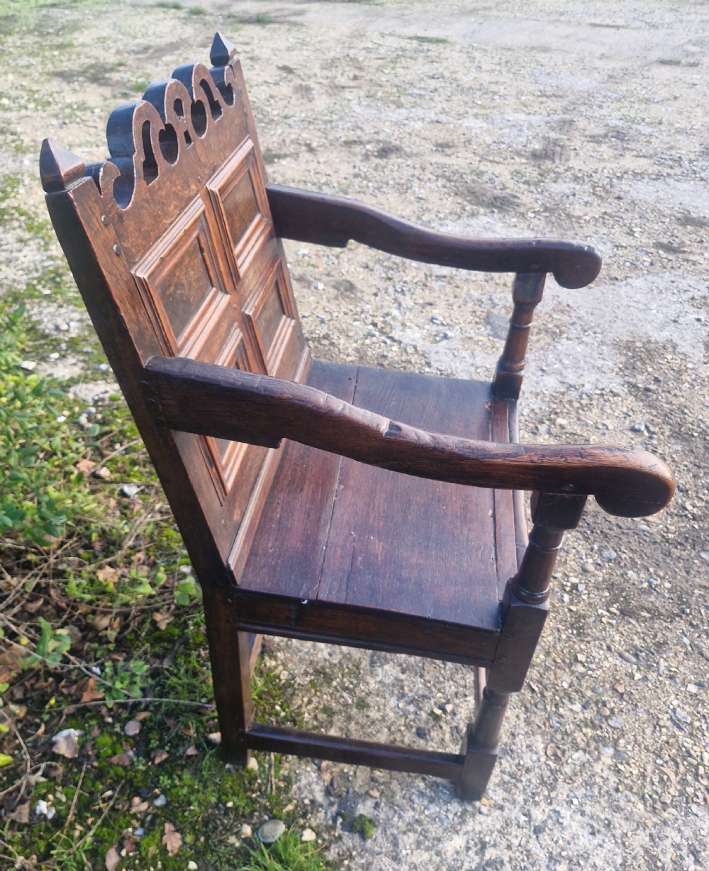 An early 18th-century dark oak wainscot armchair featuring a pierced carved cresting rail, four recessed geometric back panels, curved armrests, and turned front legs. View IX