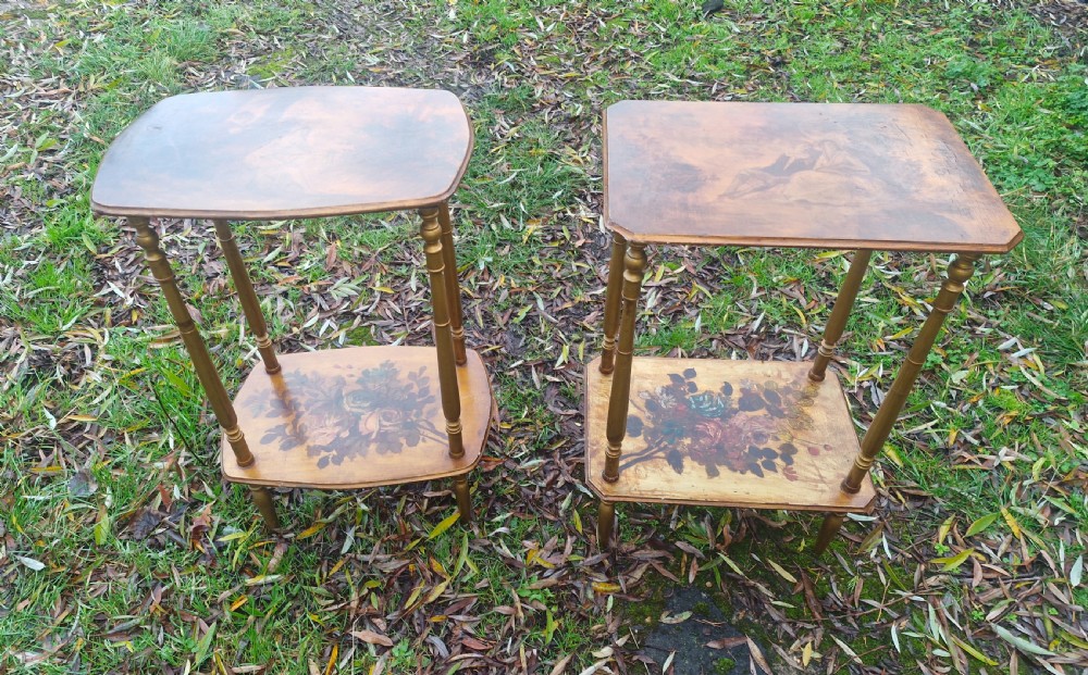 A pair of two-tier rectangular occasional tables featuring gold-painted turned legs, painted figurative scenes on the top surfaces, and floral artwork on the lower shelves.