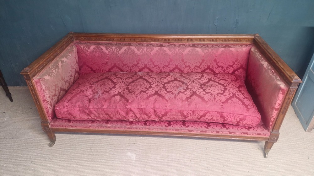 An Edwardian mahogany frame sofa upholstered in rose pink damask fabric with carved detailing along the top rail and square legs on castors.