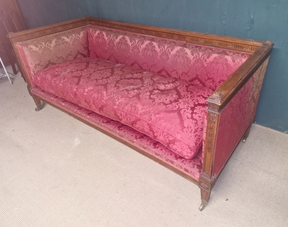 An Edwardian mahogany frame sofa upholstered in rose pink damask fabric with carved detailing along the top rail and square legs on castors. View V