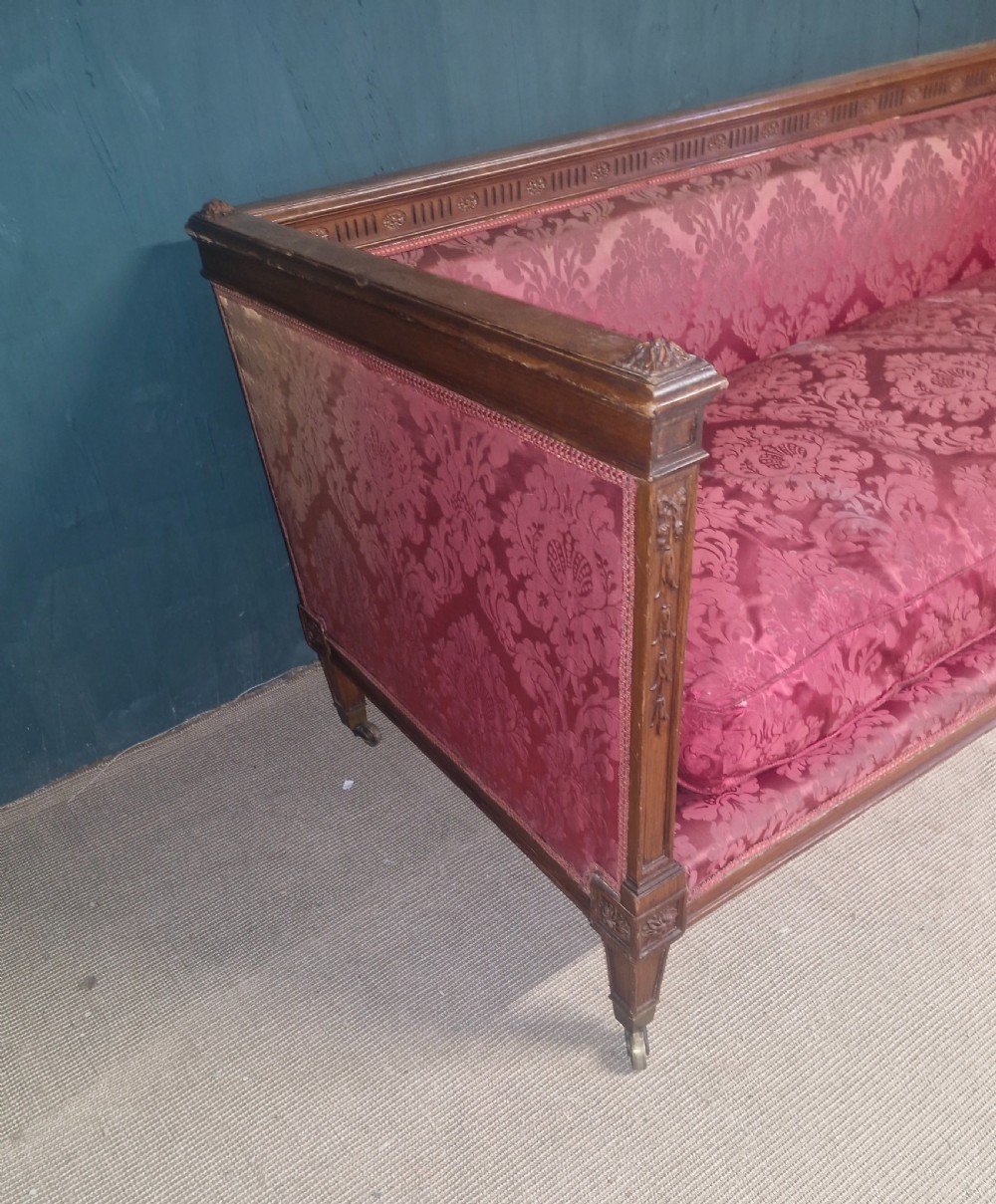 An Edwardian mahogany frame sofa upholstered in rose pink damask fabric with carved detailing along the top rail and square legs on castors. View VI
