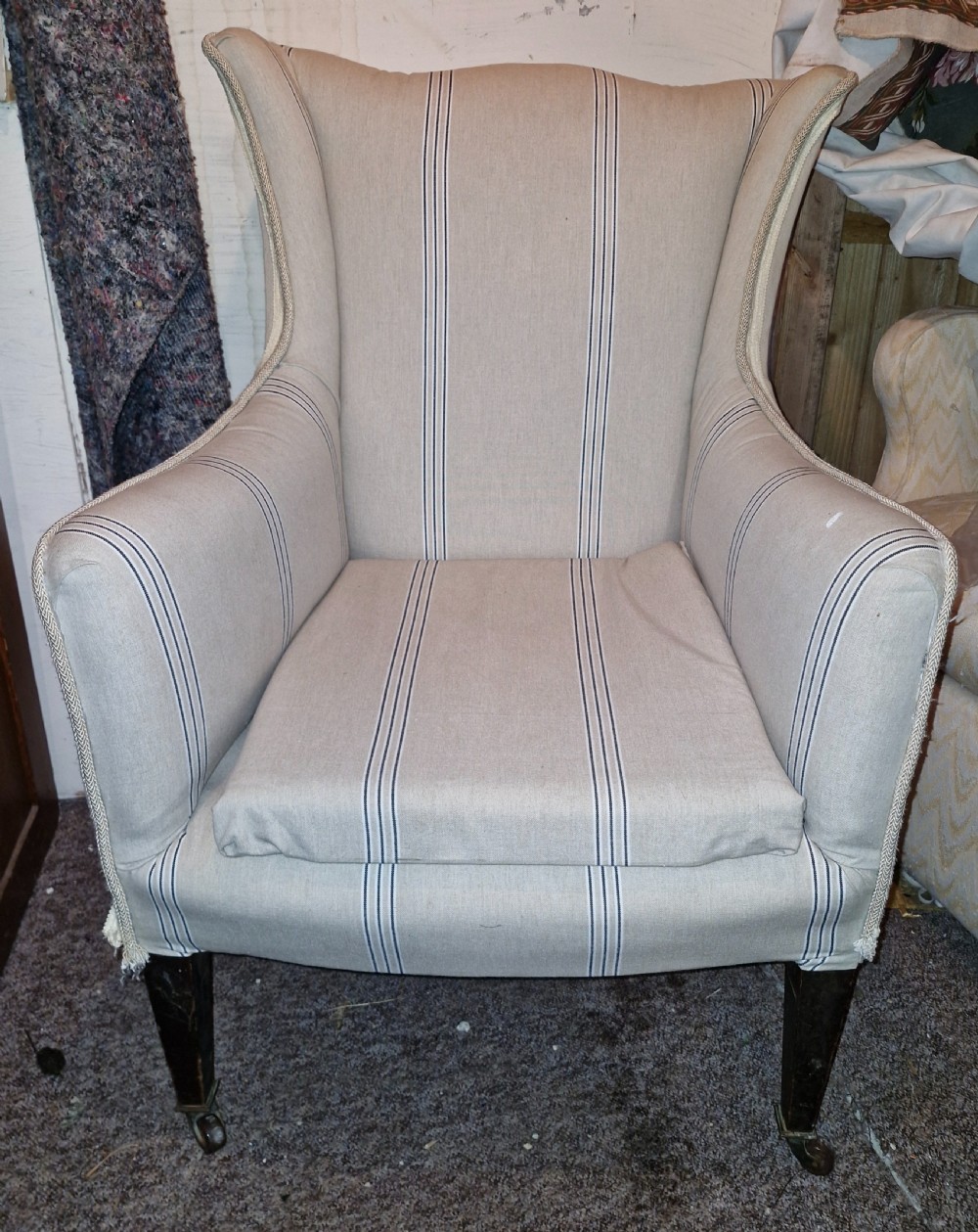 An Edwardian upholstered bedroom armchair covered in beige ticking stripe linen with tapered front legs set on brass castors.