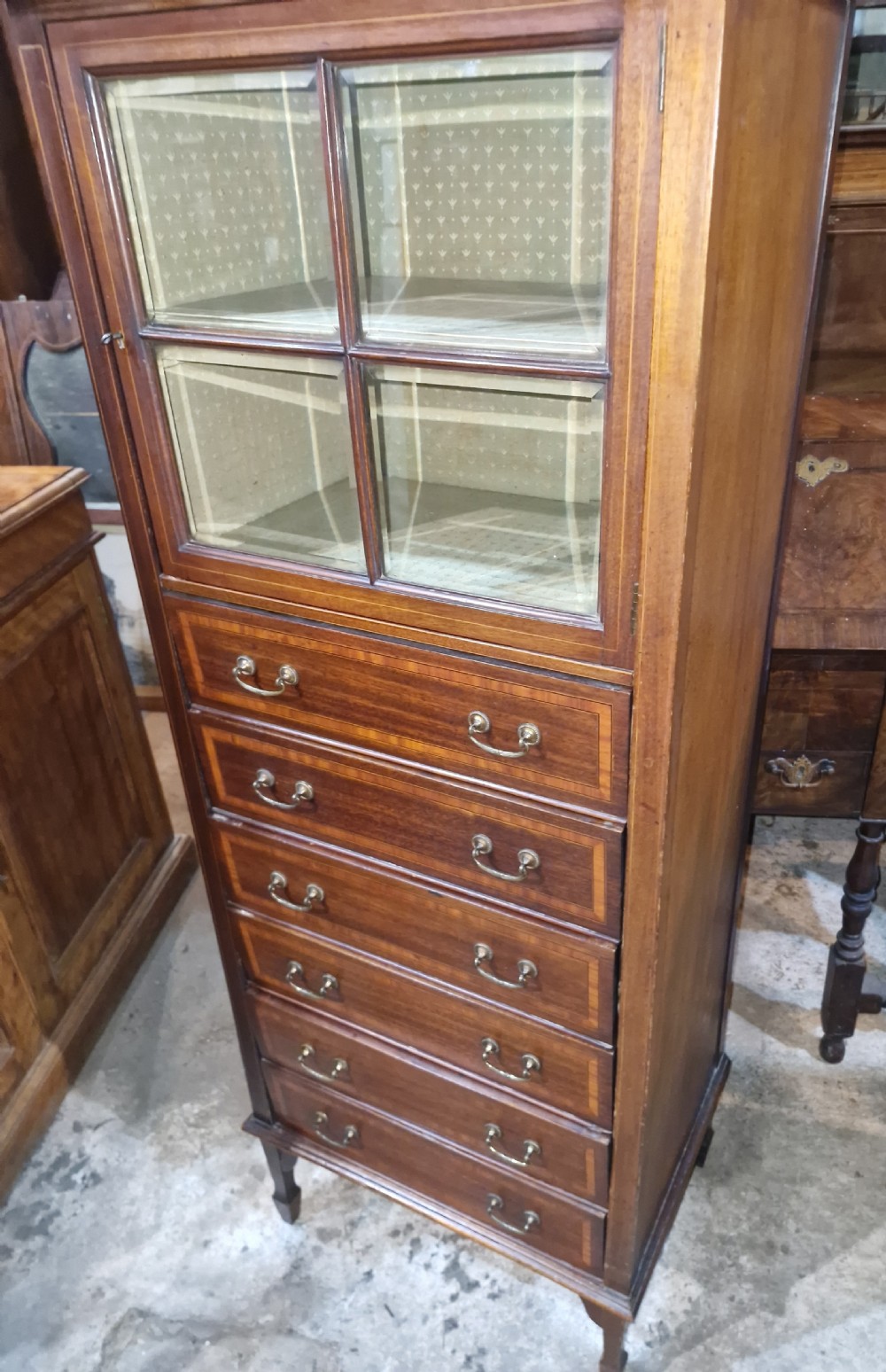 An Edwardian mahogany music cabinet featuring a four-pane glazed upper door above six inlaid drawers with brass drop handles. View VI