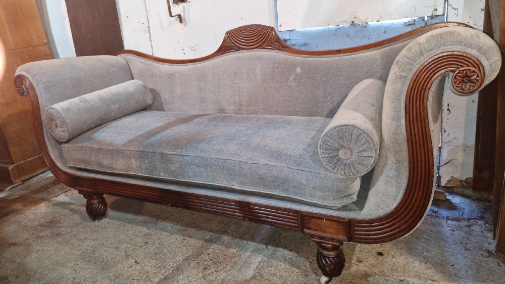 A Regency period mahogany hall sofa upholstered in grey fabric with scrolled ends, reeded frame details, shell cresting, turned spiralled feet on castors, and two cylindrical bolster cushions.