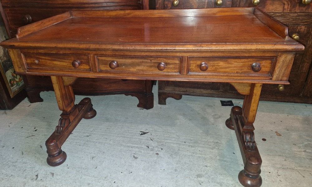 An early Victorian mahogany writing table featuring a low gallery rim, three frieze drawers with turned wooden knobs, and a trestle base with thick turned feet.