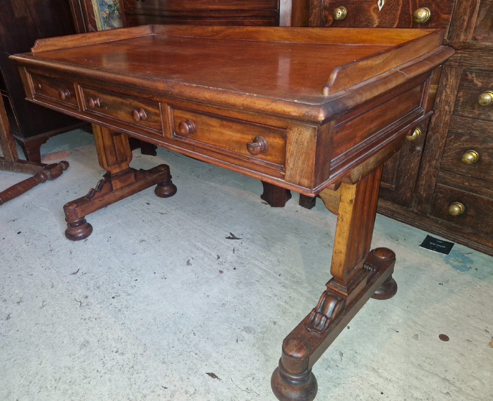 An early Victorian mahogany writing table featuring a low gallery rim, three frieze drawers with turned wooden knobs, and a trestle base with thick turned feet. View II