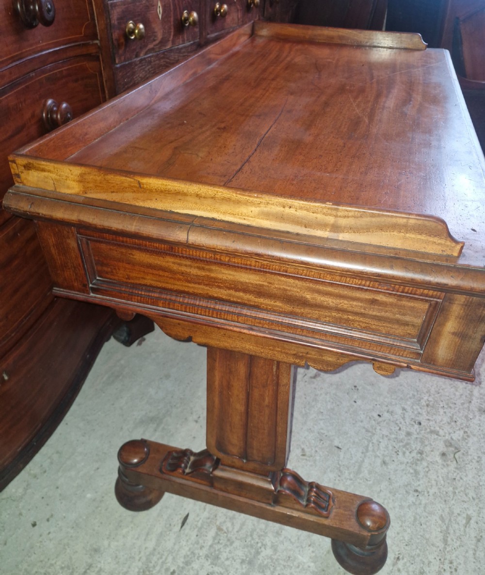 An early Victorian mahogany writing table featuring a low gallery rim, three frieze drawers with turned wooden knobs, and a trestle base with thick turned feet. View IV