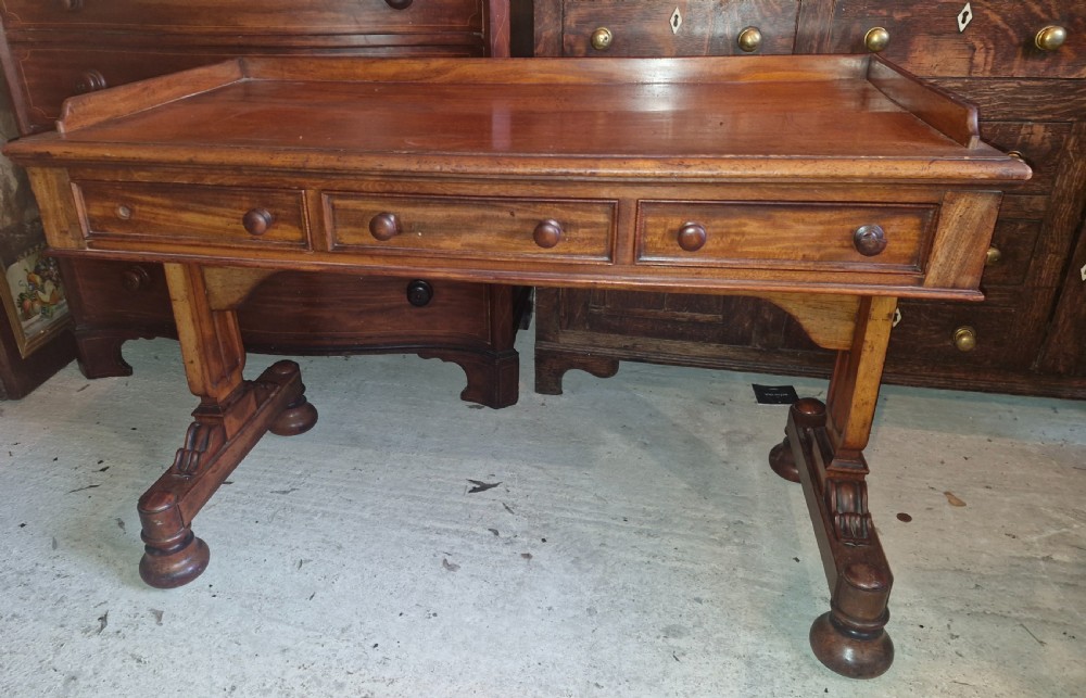 An early Victorian mahogany writing table featuring a low gallery rim, three frieze drawers with turned wooden knobs, and a trestle base with thick turned feet. View V