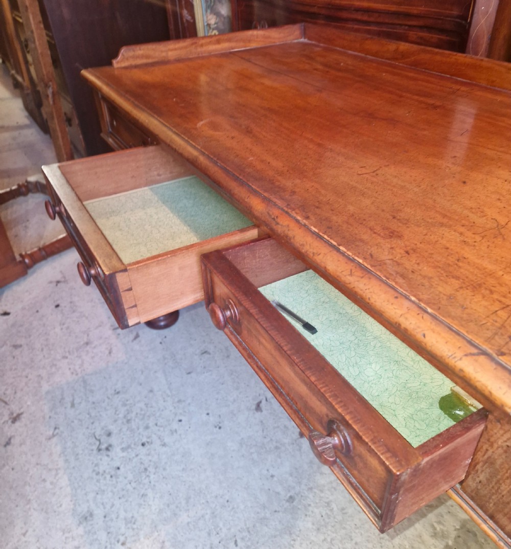An early Victorian mahogany writing table featuring a low gallery rim, three frieze drawers with turned wooden knobs, and a trestle base with thick turned feet. View VI