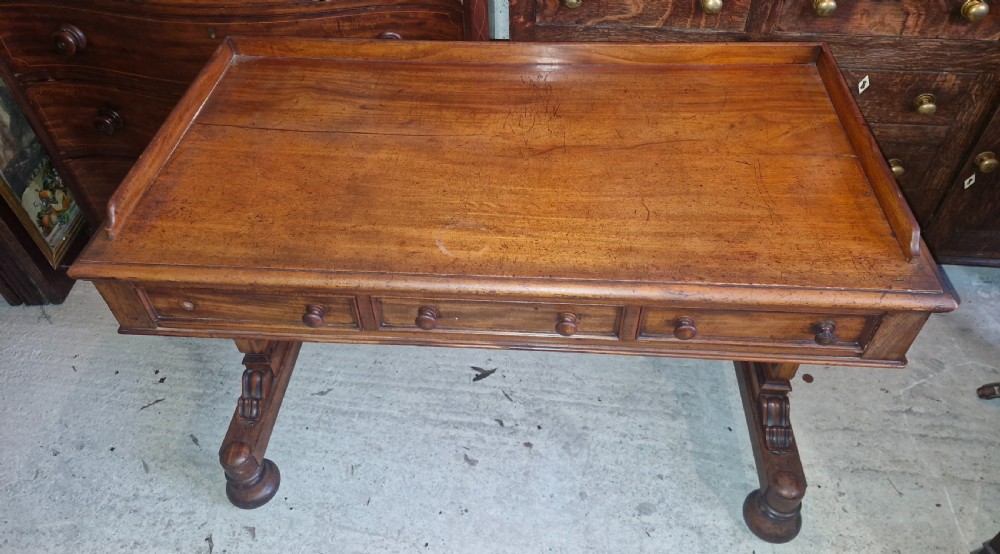 An early Victorian mahogany writing table featuring a low gallery rim, three frieze drawers with turned wooden knobs, and a trestle base with thick turned feet. View VIII
