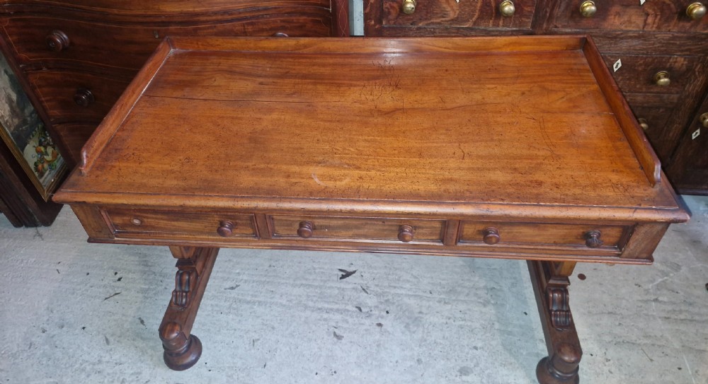 An early Victorian mahogany writing table featuring a low gallery rim, three frieze drawers with turned wooden knobs, and a trestle base with thick turned feet. View IX