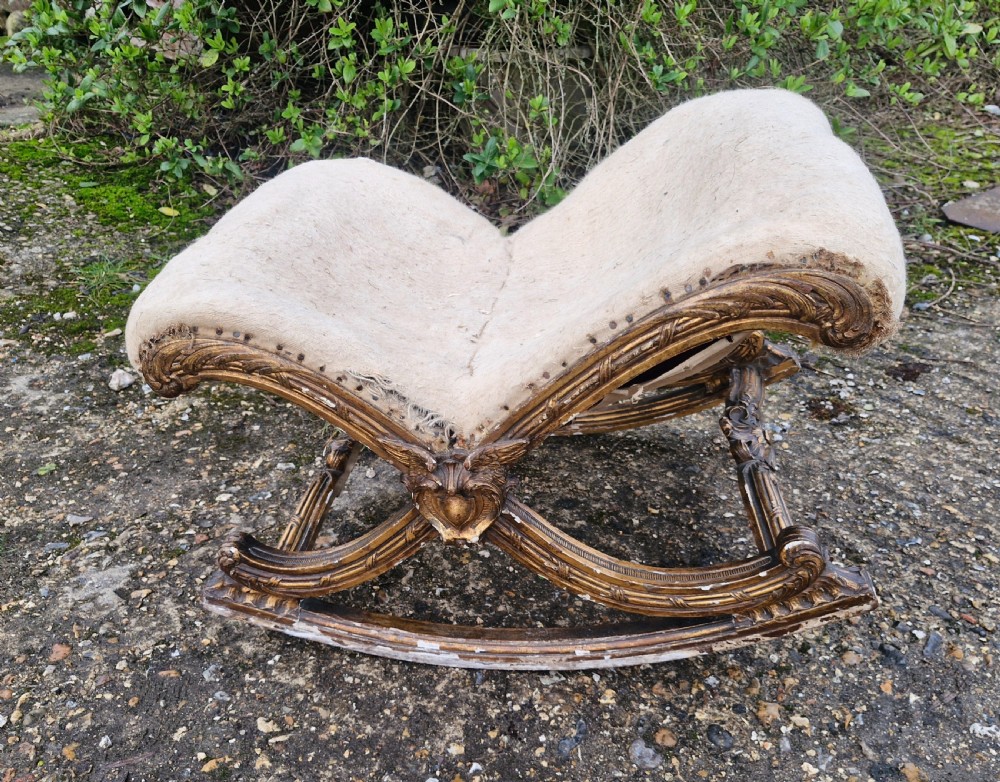 A late 19th-century giltwood rocking gout stool featuring a deeply curved cream fabric-upholstered seat supported by an ornate carved gilded frame.