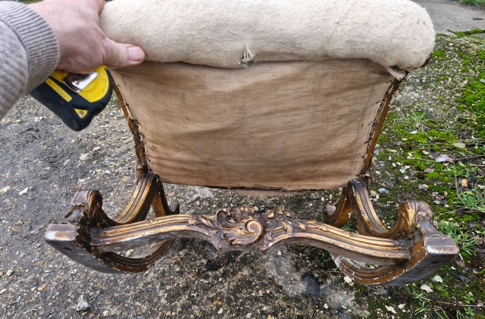 A late 19th-century giltwood rocking gout stool featuring a deeply curved cream fabric-upholstered seat supported by an ornate carved gilded frame. View V