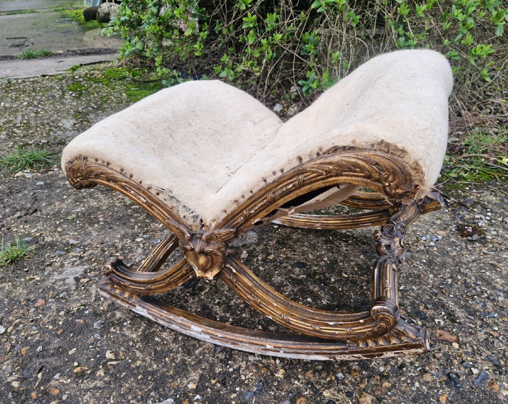A late 19th-century giltwood rocking gout stool featuring a deeply curved cream fabric-upholstered seat supported by an ornate carved gilded frame. View VI