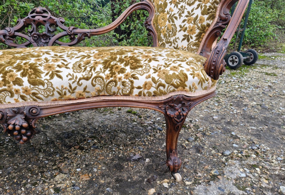 A Victorian carved walnut conversation seat featuring an S-shaped serpentine frame, floral patterned velvet upholstery, and carved cabriole legs on castors. View V