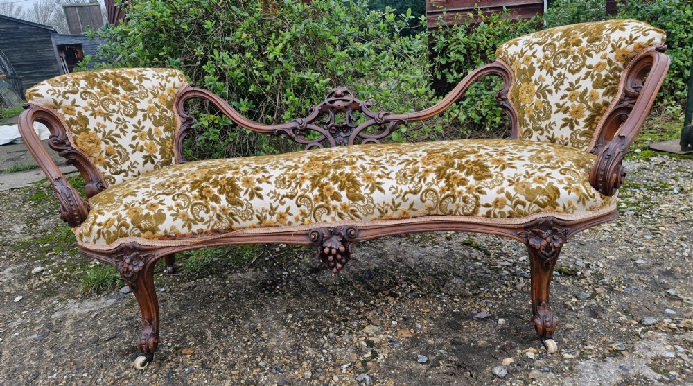 A Victorian carved walnut conversation seat featuring an S-shaped serpentine frame, floral patterned velvet upholstery, and carved cabriole legs on castors. View VI