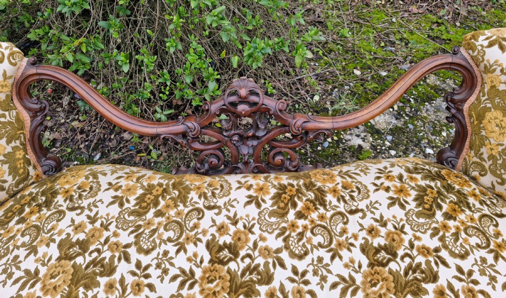 A Victorian carved walnut conversation seat featuring an S-shaped serpentine frame, floral patterned velvet upholstery, and carved cabriole legs on castors. View IX