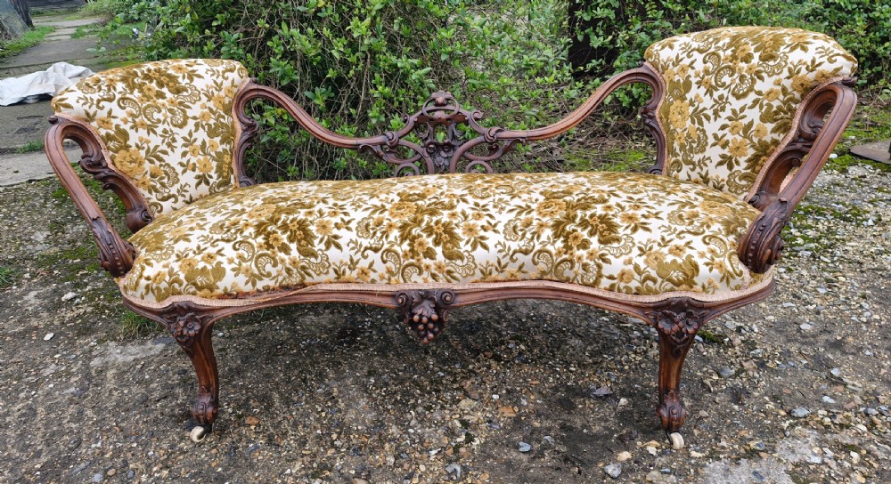 A Victorian carved walnut conversation seat featuring an S-shaped serpentine frame, floral patterned velvet upholstery, and carved cabriole legs on castors. View X