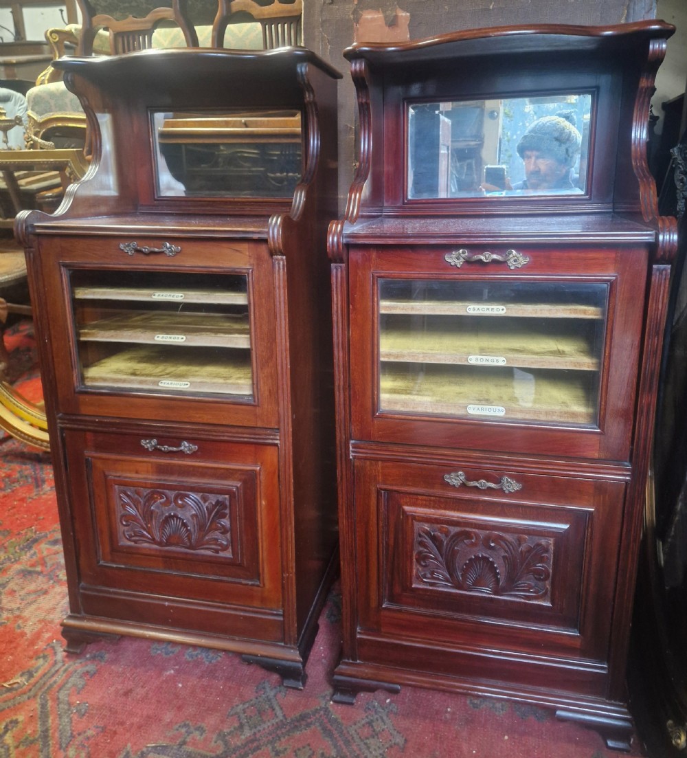 A matching pair of late Victorian dark mahogany music cabinets featuring upper mirrored shelves, glazed doors revealing labelled shelves, and lower carved wood panels.