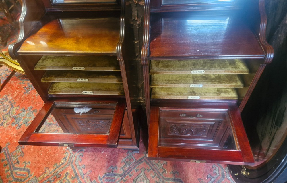 A matching pair of late Victorian dark mahogany music cabinets featuring upper mirrored shelves, glazed doors revealing labelled shelves, and lower carved wood panels. View V