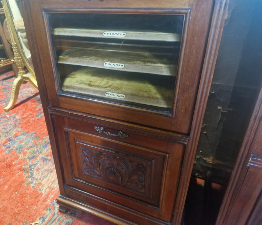 A matching pair of late Victorian dark mahogany music cabinets featuring upper mirrored shelves, glazed doors revealing labelled shelves, and lower carved wood panels. View X