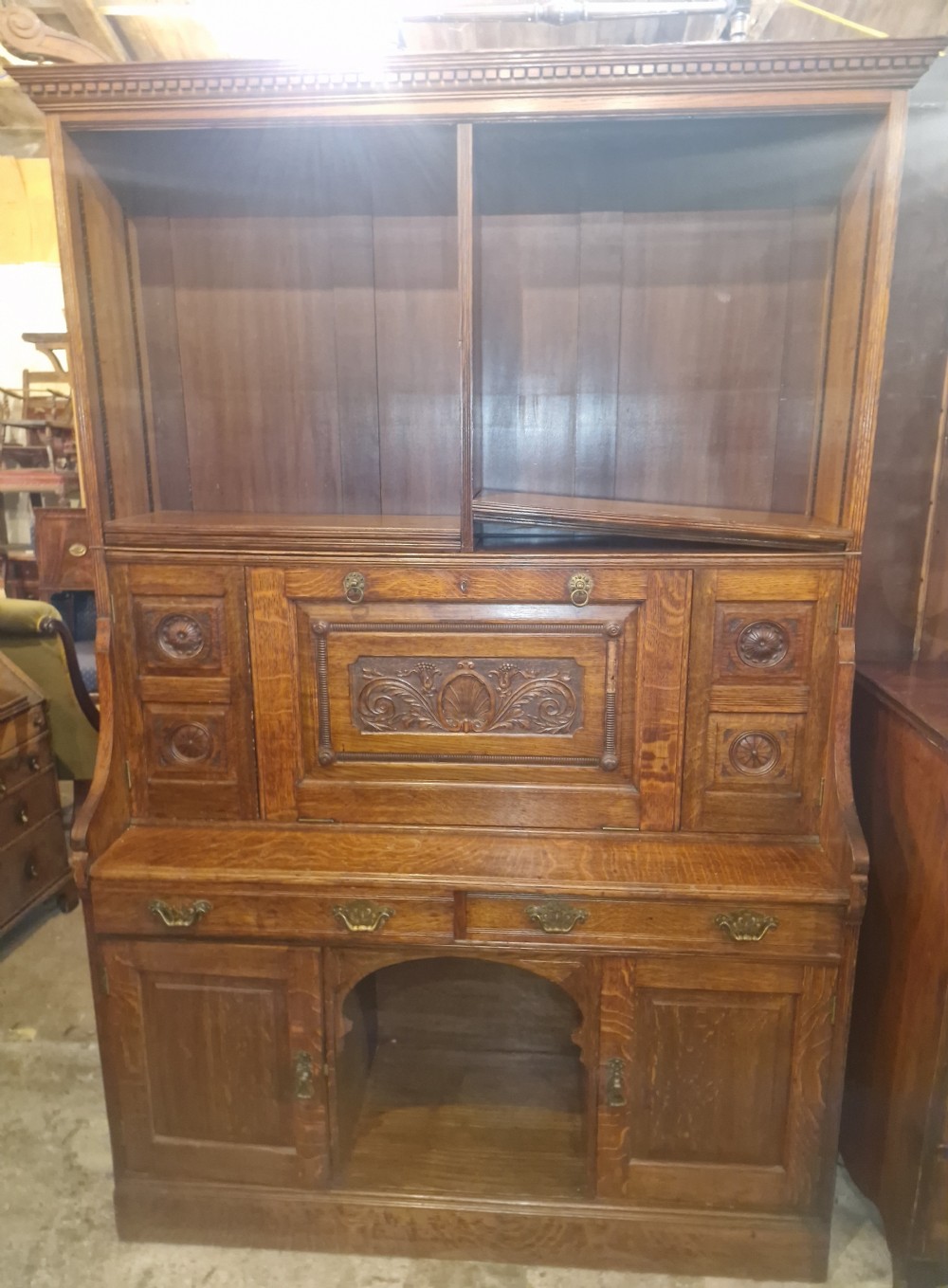 A late Victorian medium brown oak bureau bookcase with an open upper shelf unit, carved fall-front writing desk section, and lower cupboards.