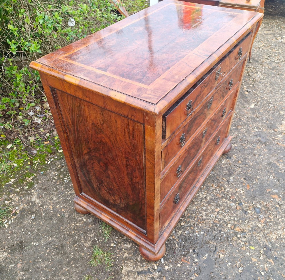 An antique walnut chest of four drawers featuring highly figured burr veneers, inlaid crossbanding, metal handles, and turned bun feet. View V