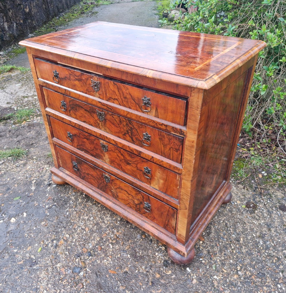 An antique walnut chest of four drawers featuring highly figured burr veneers, inlaid crossbanding, metal handles, and turned bun feet. View VI