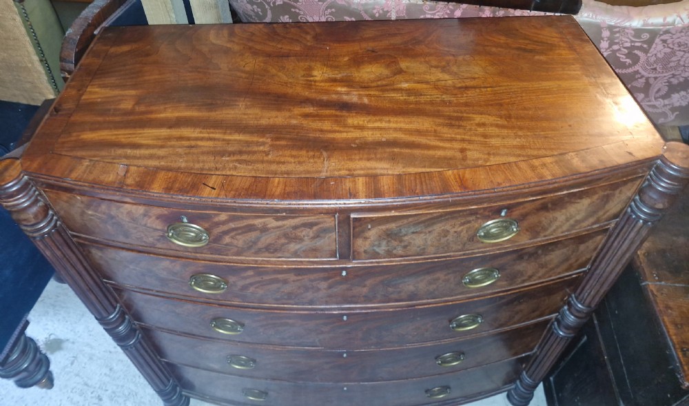 A Regency mahogany bow-front chest of drawers featuring two short over three long drawers with flame mahogany veneers, reeded side columns, and oval brass handles. View VI