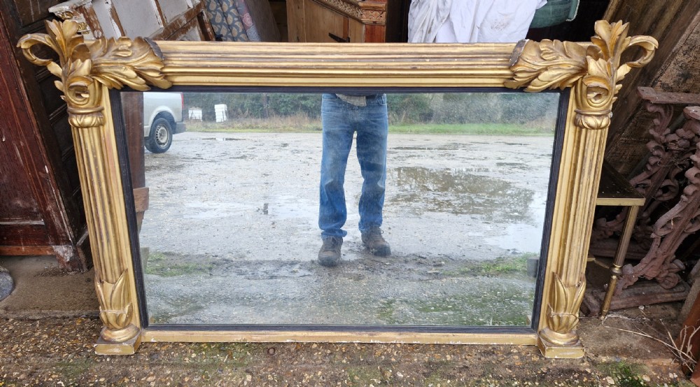 A wide rectangular Regency giltwood overmantel mirror featuring carved acanthus leaf corner details, fluted side columns, and an ebonised inner border.