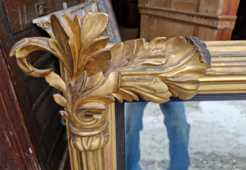 A wide rectangular Regency giltwood overmantel mirror featuring carved acanthus leaf corner details, fluted side columns, and an ebonised inner border. View V