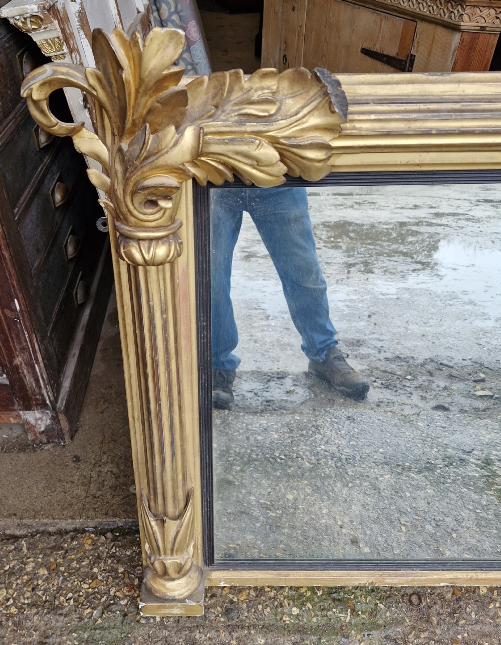 A wide rectangular Regency giltwood overmantel mirror featuring carved acanthus leaf corner details, fluted side columns, and an ebonised inner border. View VI