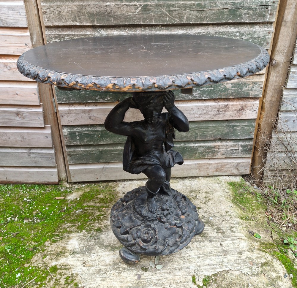A carved ebonised wooden occasional table featuring a figural pedestal base supporting a circular top with a leaf-carved border. View II
