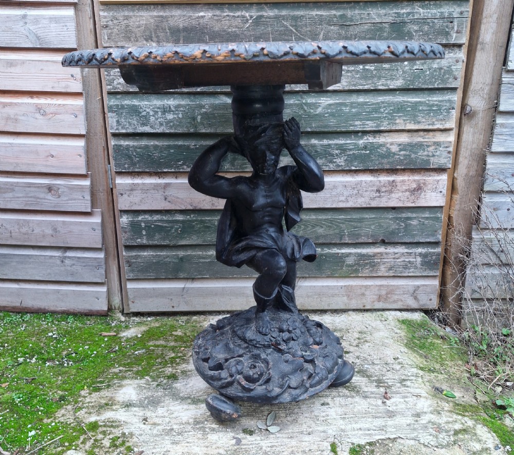 A carved ebonised wooden occasional table featuring a figural pedestal base supporting a circular top with a leaf-carved border. View IV
