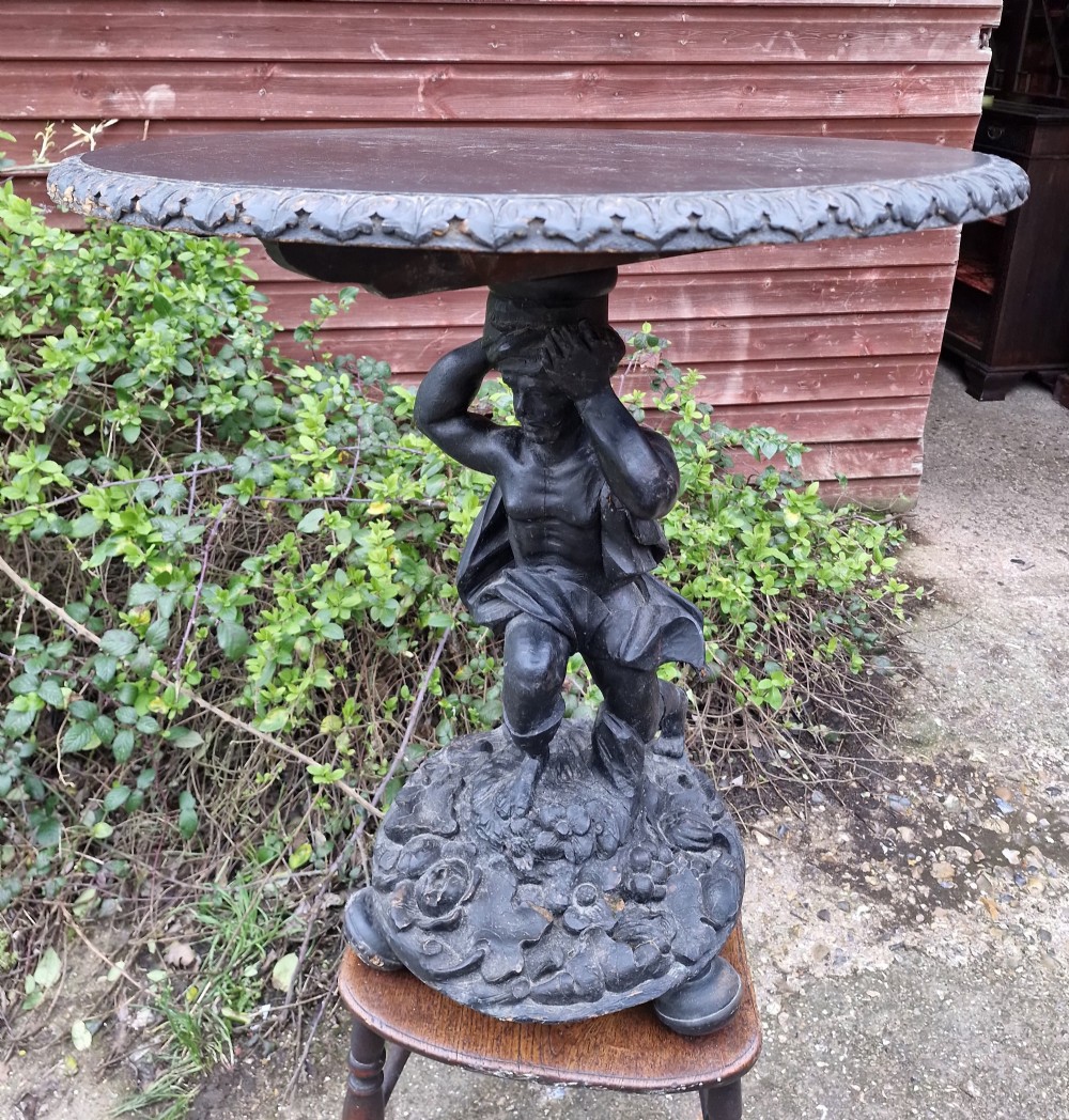 A carved ebonised wooden occasional table featuring a figural pedestal base supporting a circular top with a leaf-carved border.