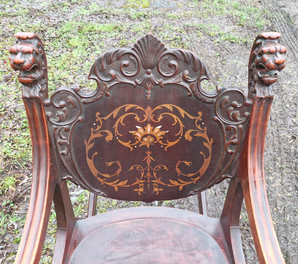 A late 19th-century dark mahogany rocking armchair featuring carved lion head uprights and a floral inlaid backrest. View V