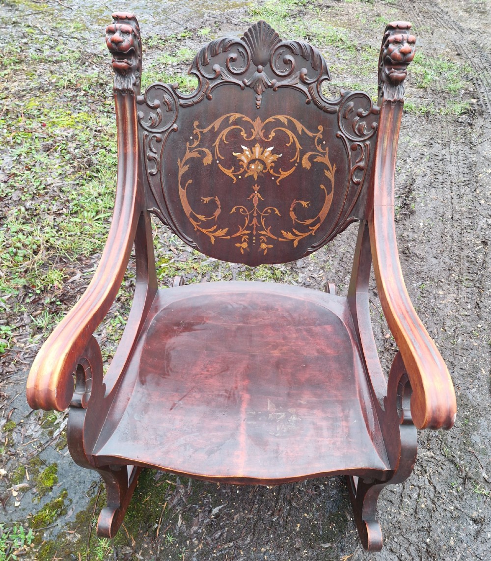 A late 19th-century dark mahogany rocking armchair featuring carved lion head uprights and a floral inlaid backrest. View VI