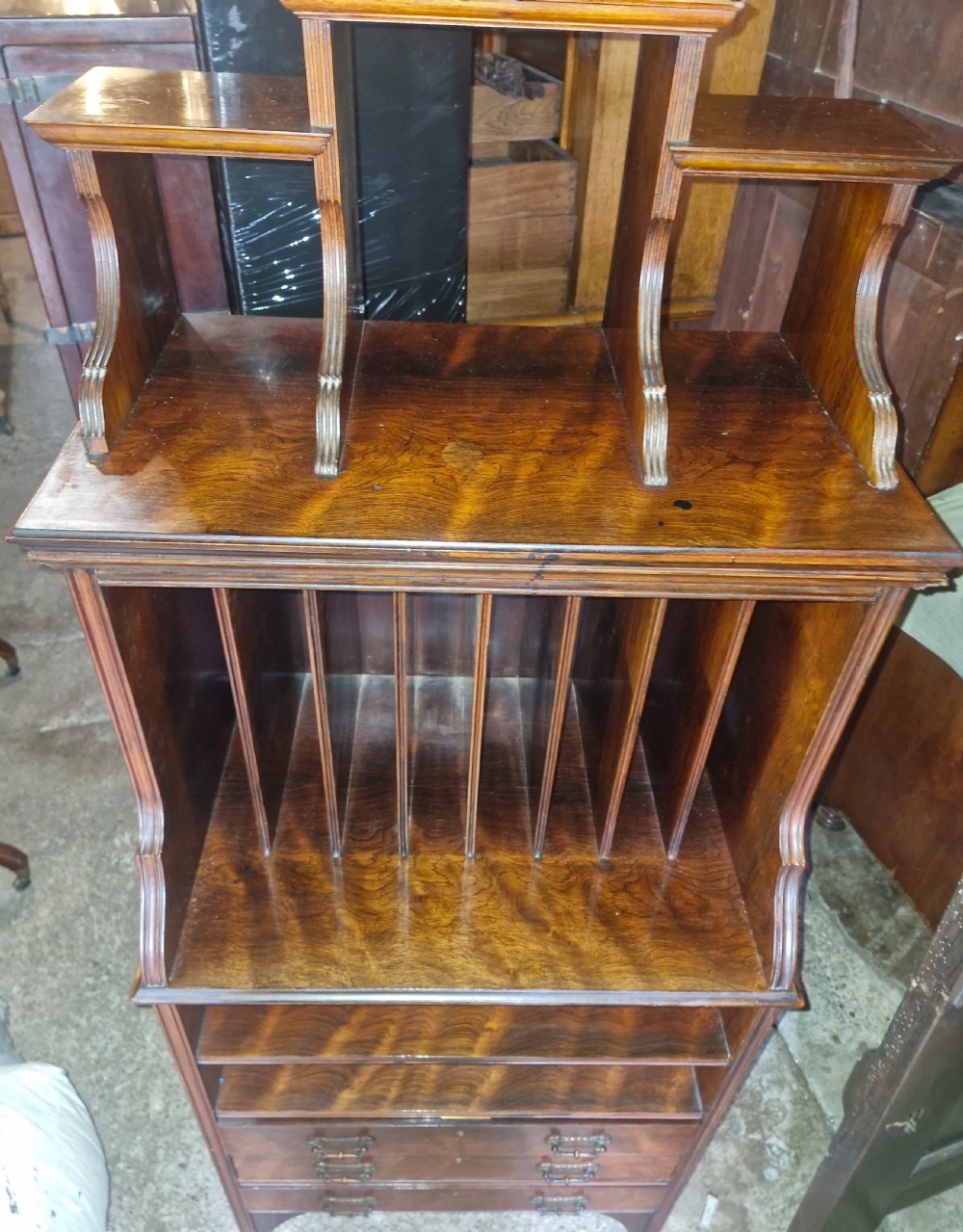 A late Victorian dark rosewood side cabinet featuring upper tiered display shelves, vertical open folio partitions, lower open shelving, and small drawers with brass drop handles. View II