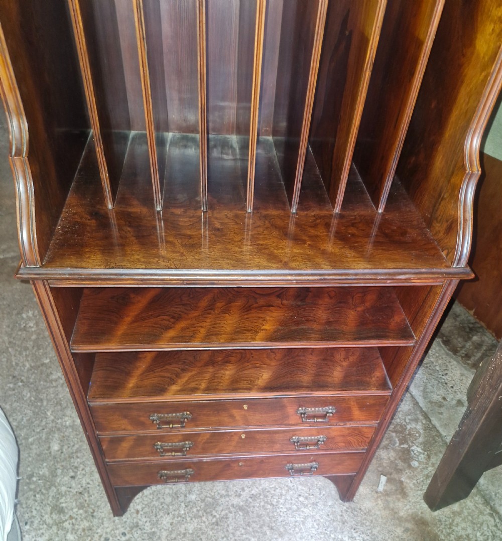 A late Victorian dark rosewood side cabinet featuring upper tiered display shelves, vertical open folio partitions, lower open shelving, and small drawers with brass drop handles. View IV